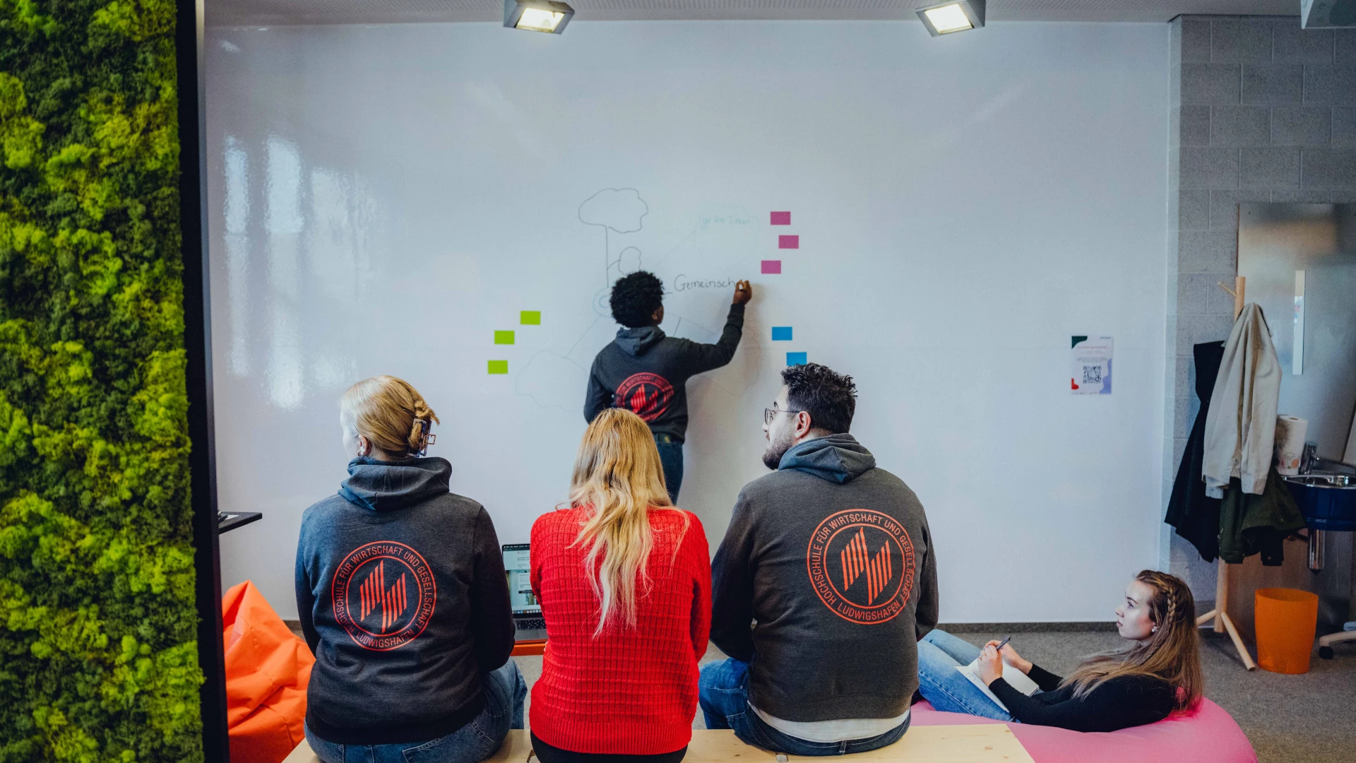 Rear view of three students sitting on a bench; one student is writing on a whiteboard