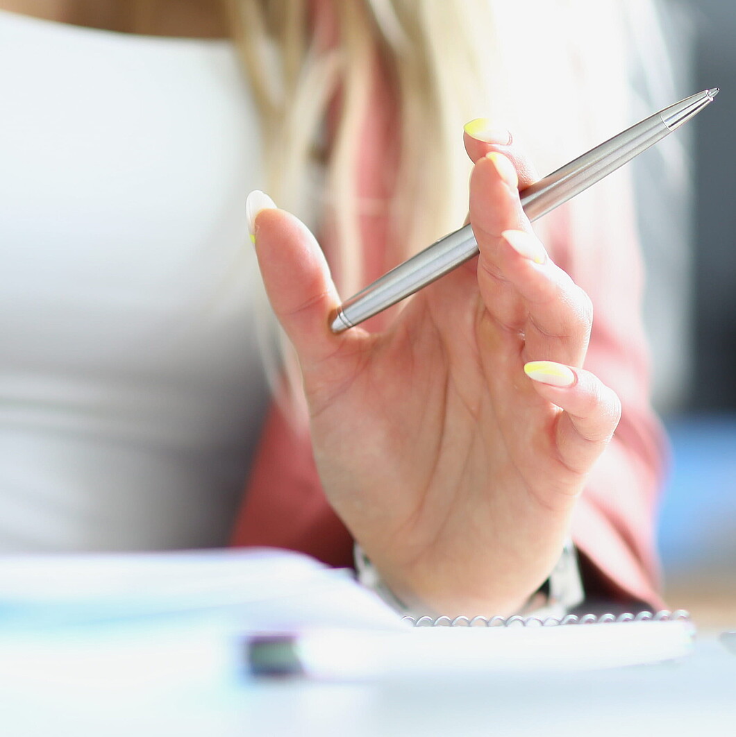 Nahaufnahme einer Person, die einen Stift hält; im Vordergrund ein Laptop, daneben ein Stapel Bücher und Notizhefte.