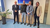 3 ältere Männer und ein Mann mittleren Alters stehen lächelnd in einem Büro
