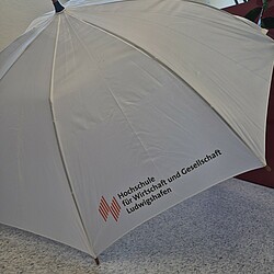 A white stick umbrella with a wooden handle and the logo of the Ludwigshafen University of Business and Society is placed open on the floor. A window with a blue sky can be seen in the background.