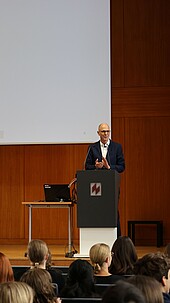 Ein Mann spricht vor Publikum an einem Podium in einem Hörsaal.