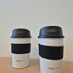 Two white thermo mugs with black lids and rubberized handles stand next to each other on a table. Both mugs bear the university logo in small red lettering.