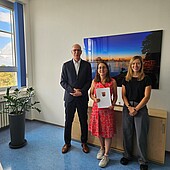A middle-aged man and two women are standing in an office, smiling. The woman in the middle is holding a certificate in her hands
