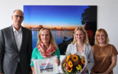 Ein älterer Mann, eine ältere Frau mit Bilderrahmen und zwei Frauen mittleren Alters mit Blumenstrauß  stehen lächelnd in einem Büro.