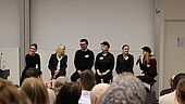 zwei Frauen und vier Studieende szuen auf Barhockern im Auditorium der Hochschule. Eine Person spricht gerade ins Mikrofon