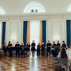 The university choir stands in a row in front of a seated audience in a festive room with high windows and blue and gold curtains and sings together.