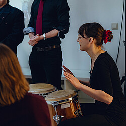 A young woman sits at the side of a wall and plays two congas in concentration. Listeners can be seen in the foreground.