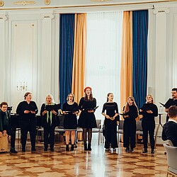 The university choir sings in a row in front of a seated audience in a festive hall. Conga drums and a cajón can be seen on the left, played by a choir member. The singers are wearing black clothing, while one singer is wearing a red beret.