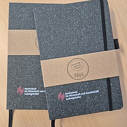Two black notebooks with a textured surface and an elastic band lie on a wooden table. The logo of the Ludwigshafen University of Business and Society is printed on both covers.