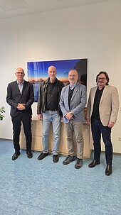 3 ältere Männer und ein Mann mittleren Alters stehen lächelnd in einem Büro
