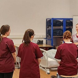 Studierende bei einer Übung a Krankenbett, zu sehen sind mehrer Studierende die im Halkreistehen und einer Übung zusehen.