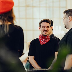 Close-up of a male choir singer smiling and wearing a red scarf while interacting with other choir members.