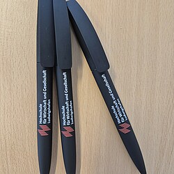 Three black ballpoint pens with a soft-touch surface and the Ludwigshafen University of Business and Society logo lie fan-shaped on a table. The pens have a simple, modern look.