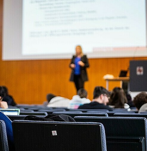 View from behind over heads in rows of chairs to a teacher in front of a large screen