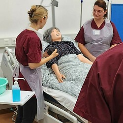 Studierende bei einer Übung a Krankenbett, zu sehen zwei Studierende mit Kittel am Bett. Im bett liegt eine Übungspuppe.