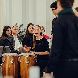The choir sings in a festive hall while a woman nearby beats the drums in the Tagt.