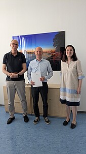 Zwei Männer und eine Frau mittleren Alters stehen lächelnd nebeneinander in einem Büro
