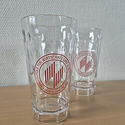 Two transparent plastic glasses with red Dubbeglas dots and the round university logo stand on a table. They are stackable and have a textured surface.