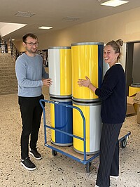 Two people stand in a hallway holding four colorful waste separation bins in yellow, blue and gray, which are placed on a blue cart. Both are smiling at the camera