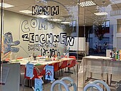 Die Fensterfront des SoIL ist bemalt und lädt zum Comiczeichnen-Workshop ein; im Hintergrund sieht man Arbeitstische und Bastelmaterial