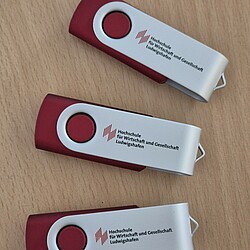 Three red and white USB sticks with a rotating mechanism and the university logo lie on a wooden table. The sticks bear the inscription "Ludwigshafen University of Business and Society".