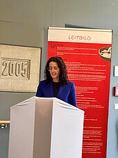 A woman is speaking to an audience; behind her is a banner with the Caritas logo.