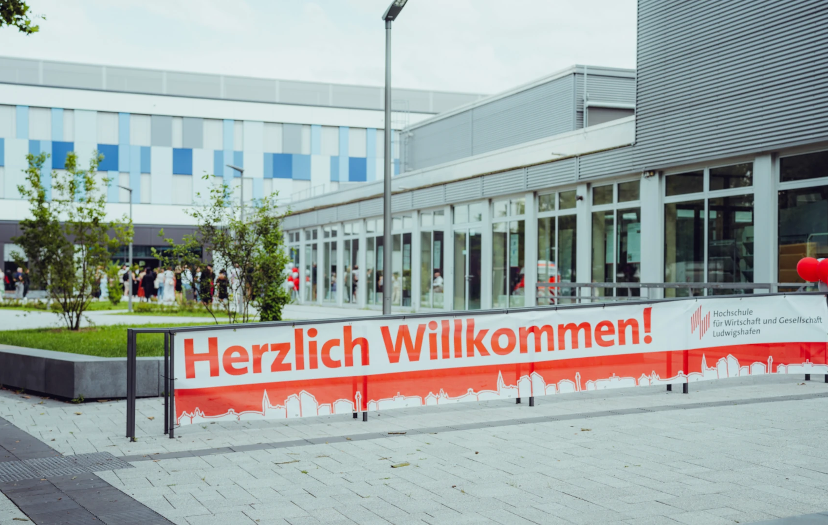banner-willkommen-vor-gebäude_hwg-lu Willkommensbanner vor modernem Gebäude, Menschen in der Nähe.