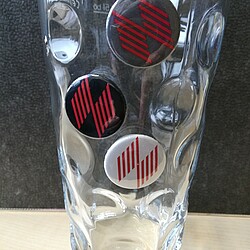 A classic dubbe glass made of glass stands on a table and contains three round buttons with the logo of the Ludwigshafen University of Business and Society. The buttons in black, gray and white are decoratively placed on the indentations of the glass.
