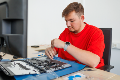 Eine Person (Auszubildende*r) arbeitet mit Schraubendreher an einem geöffneten Laptop auf einer blauen Unterlage. Daneben steht ein Monitor auf dem Schreibtisch in einem hellen Büroraum.