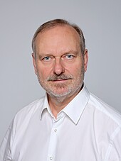 Porträtbild eines älteren Mannes mt grau melierten Haaren und Bart im weißen Hemd