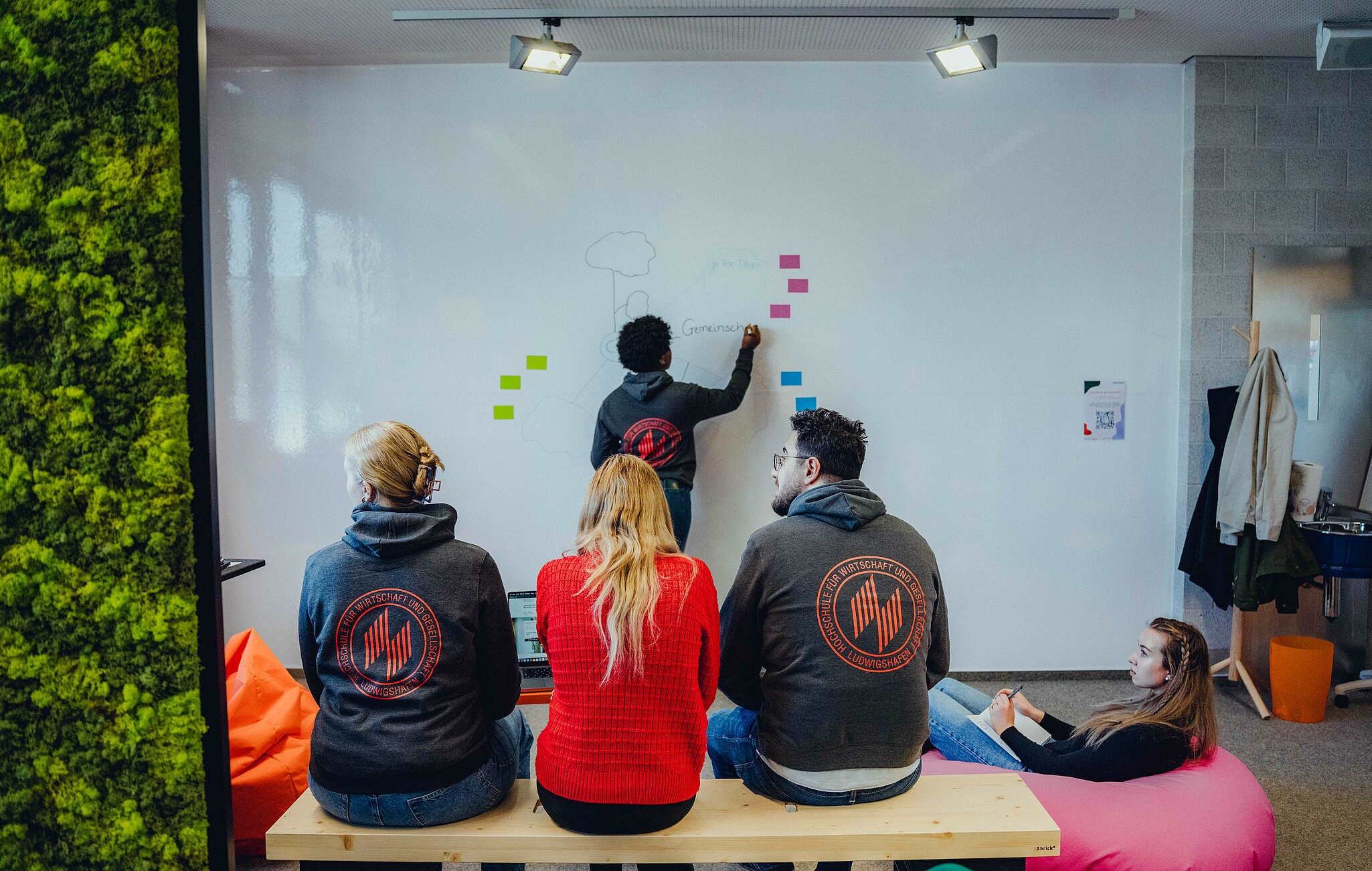 Rückenansicht von drei Studierenden auf einer Bank, eine Studierende schreibt auf ein Whiteboard