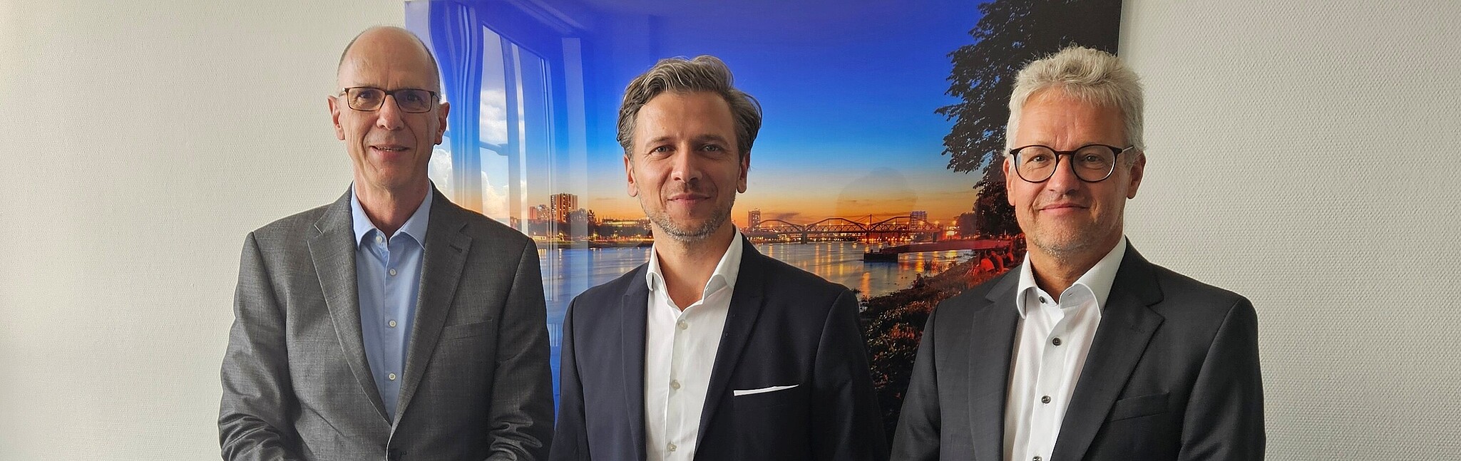 3 Männer stehen lächelnd nebeneinander in einem Büro, der mittlere Mann hält eine Zrkunde in den Händen