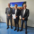 3 Männer stehen lächelnd nebeneinander in einem Büro, der mittlere Mann hält eine Zrkunde in den Händen