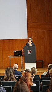 Eine Frau spricht vor einem Publikum an einem Podium in einem Hörsaal.