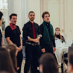 A group of choristers, including three men with colorful ties and scarves, sing together in front of the audience.