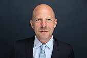 Mann mittleren Alters mit Glatze und grau meliertem Bart im Anzug mit Krawatte vor dunkelgrauem Hintergrund
