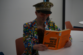 Student sitzt verkleidet in der Bibliothek und hält ein Buch mit dem Titel "Sexualerziehung in der Praxis" in der Hand