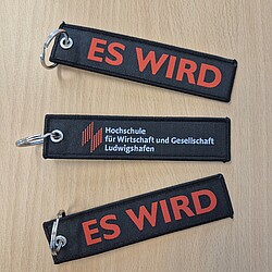 Three black textile key fobs with a metal ring are lying on a table, two show the imprint "ES WIRD" in red lettering, one also bears the university logo. The tags look sturdy and have a high-quality finish.