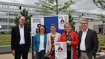 zeig_dein_gesicht_für_demokratie_hochschulleitung-HWG-2025 Zwei Männer und drei Frauen mittleren Alters stehen lächelnd auf dem Campus vor Plakaten der Fotoaktion "Zeig Dein Gesicht für Demokratie"