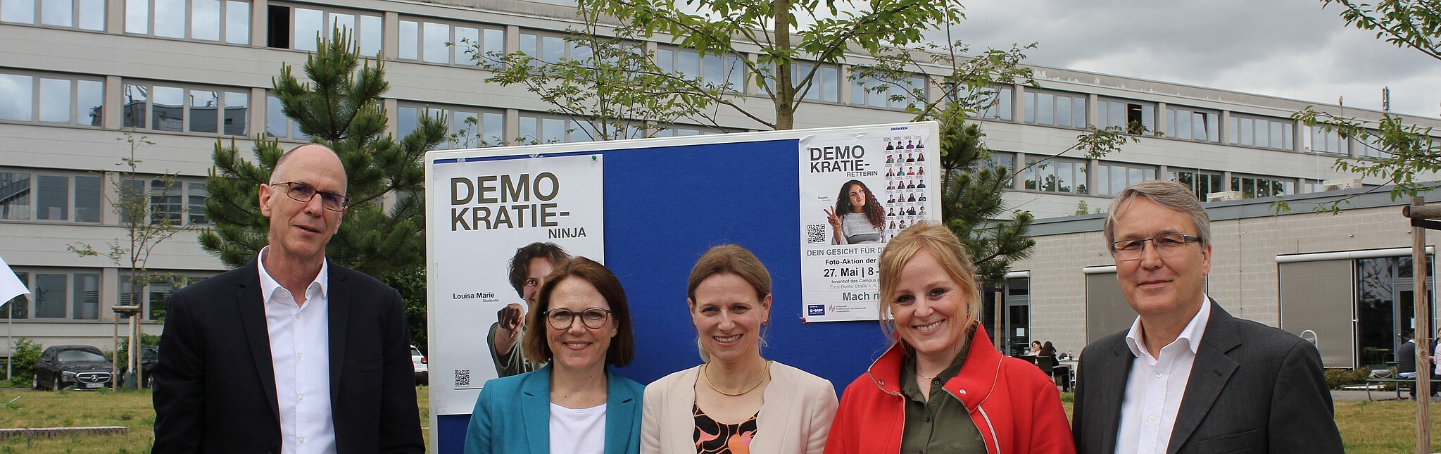 Zwei Männer und drei Frauen mittleren Alters stehen lächelnd auf dem Campus vor Plakaten der Fotoaktion "Zeig Dein Gesicht für Demokratie"