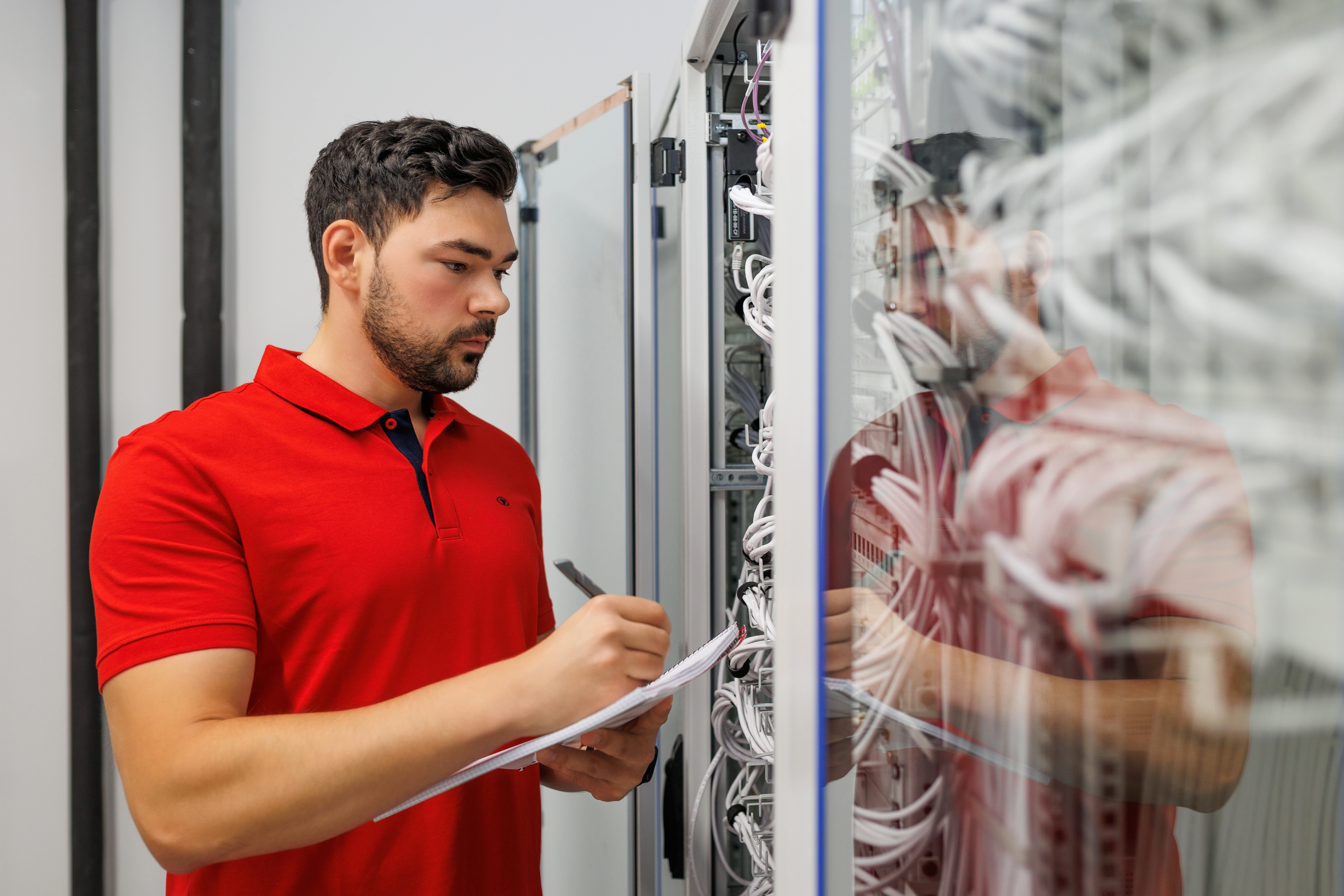 Eine Person (Auszubildende*r) steht am geöffneten Netzwerkschrank und notiert etwas auf einem Klemmbrett. In den Schränken sind viele weiße Patchkabel zu sehen, die Person spiegelt sich in der Glastür.